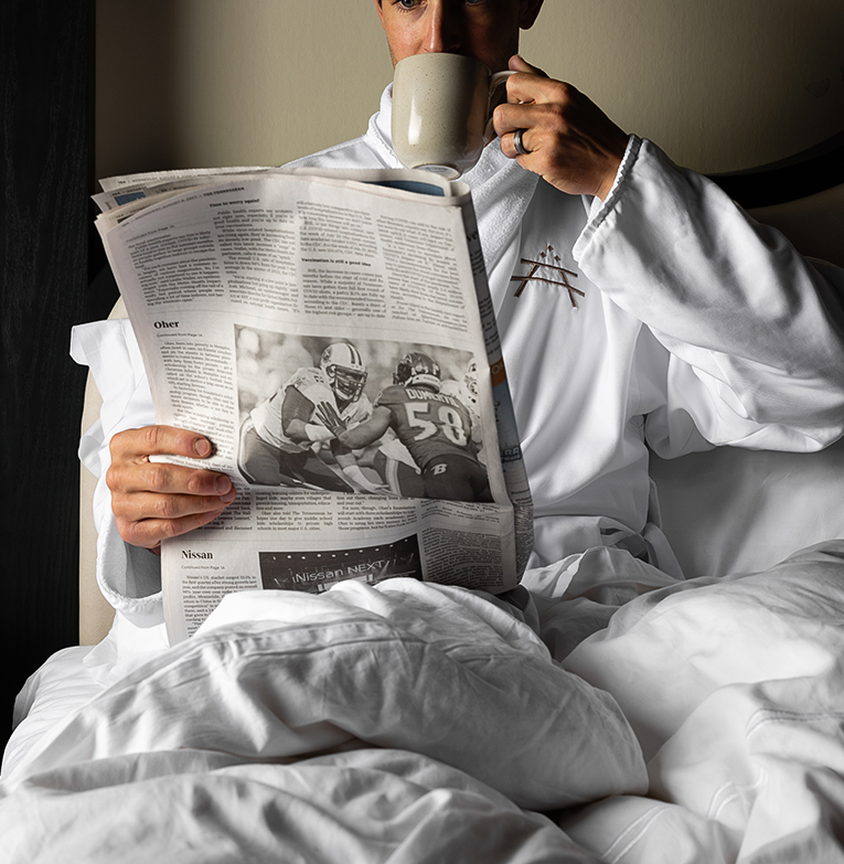a man in bed reading a newspaper and drinking coffee