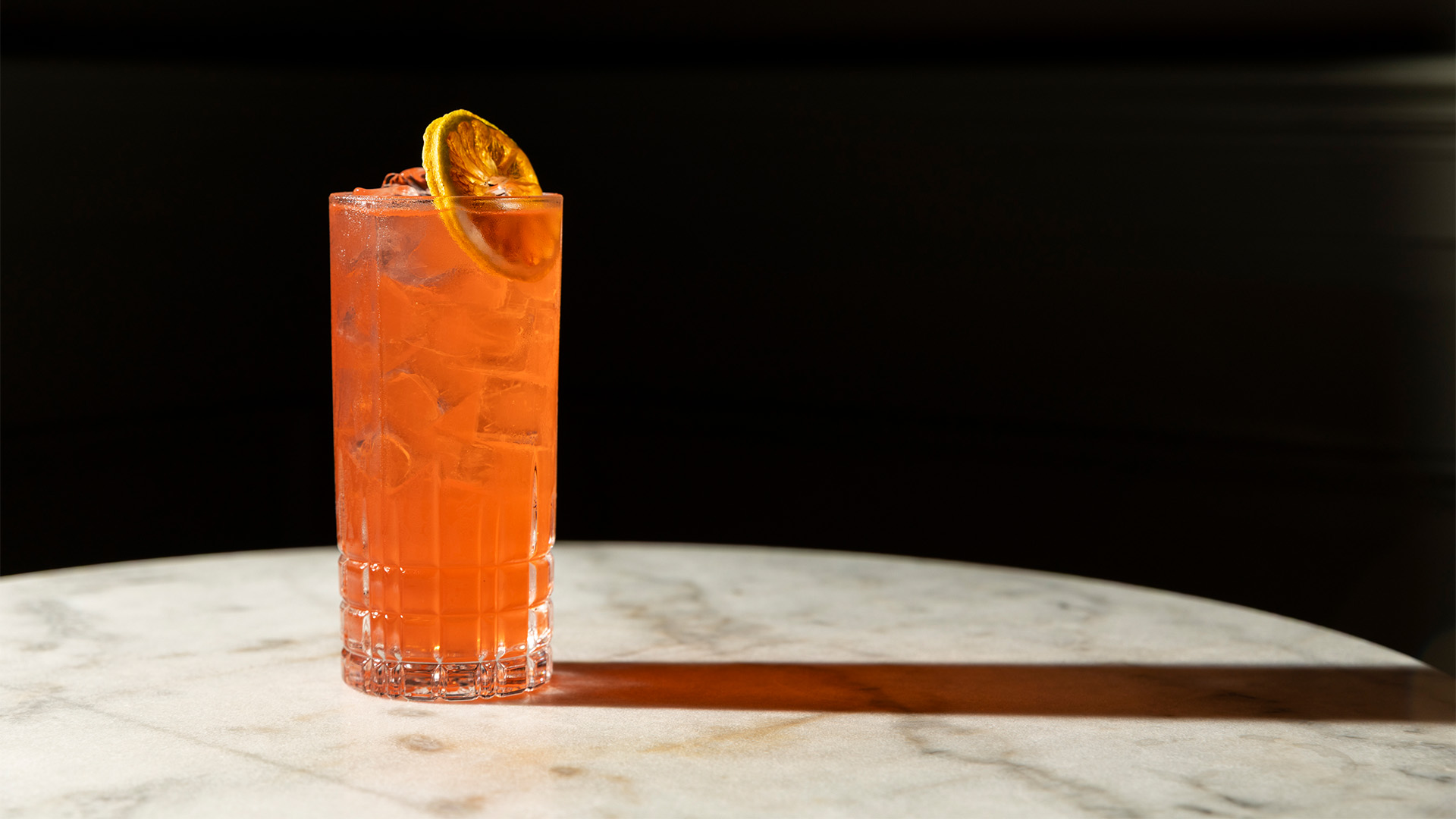 a glass of orange drink with ice and a slice of orange on a marble table