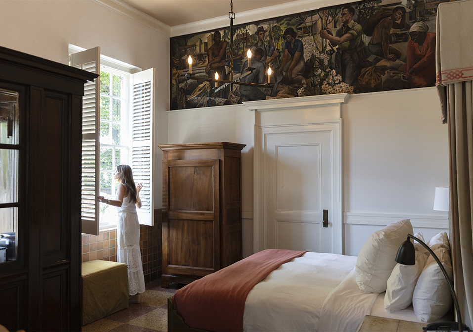 a woman standing in a room with a bed and a painting above her