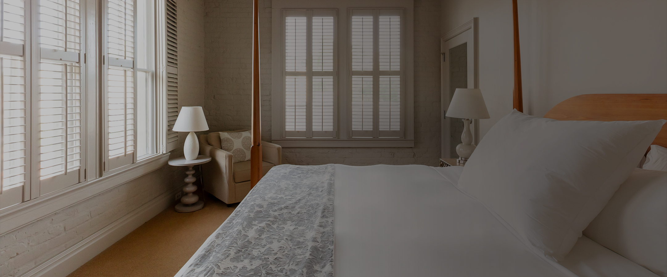 a bed with a white sheet and a lamp in a room with windows