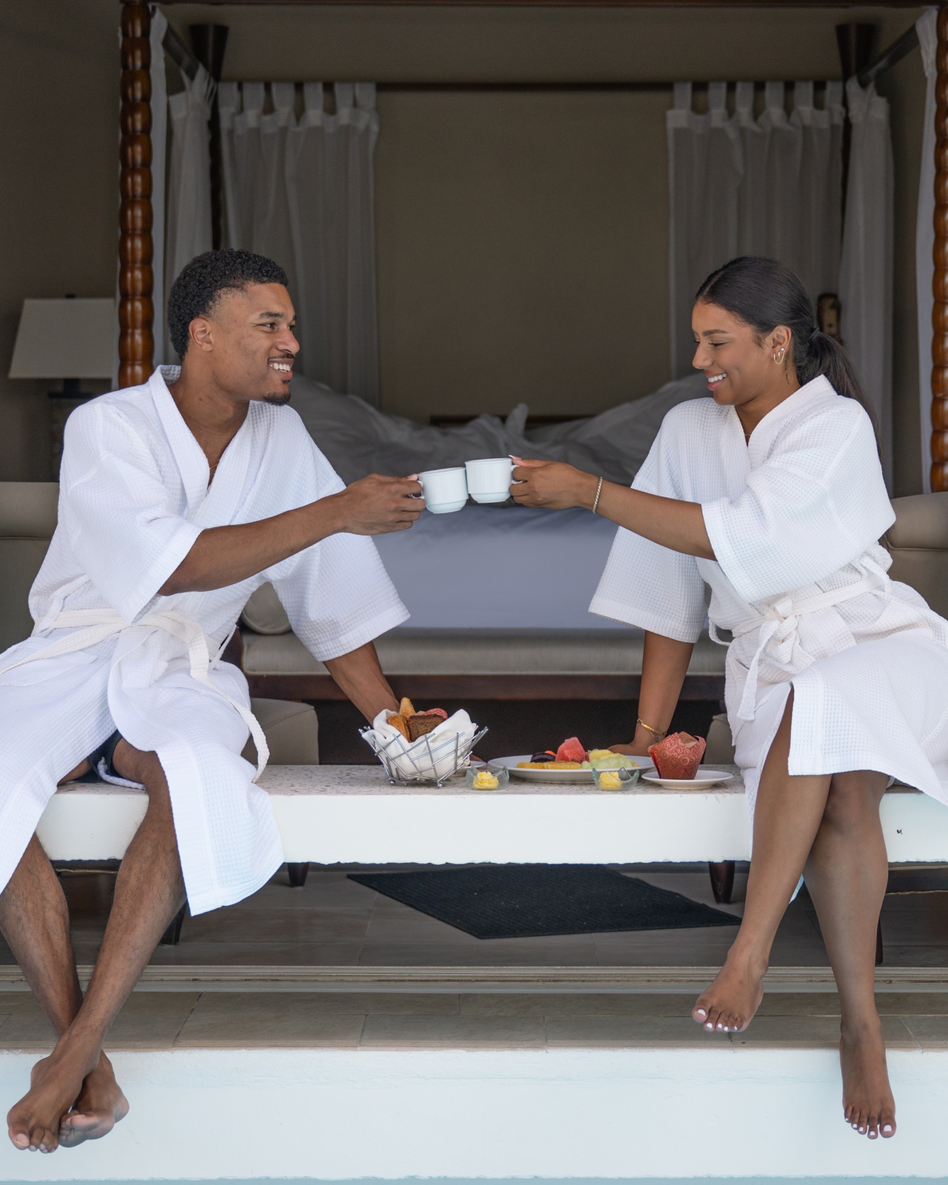 a man and woman in white robes sitting on a bed with cups of coffee