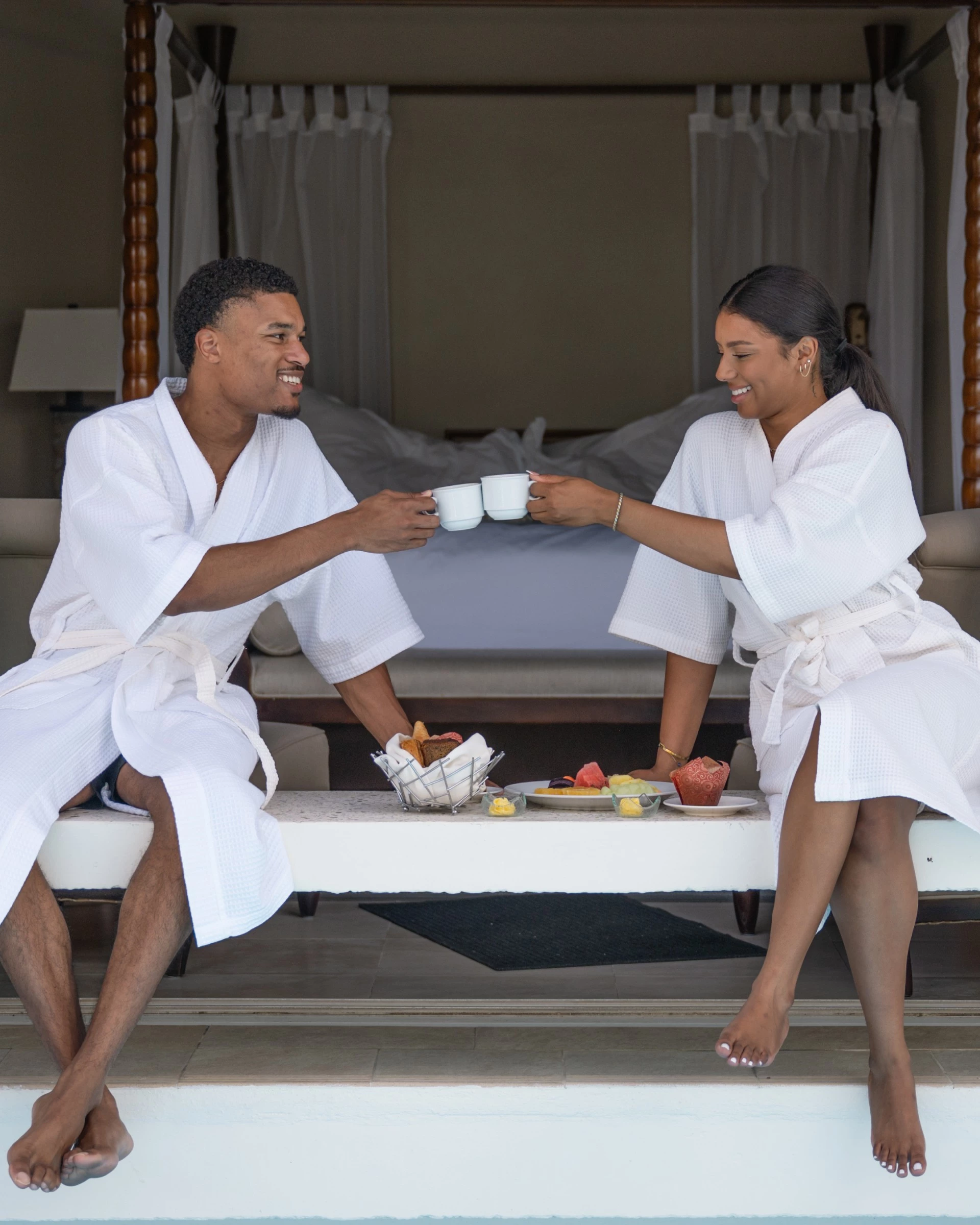 a man and woman in white robes sitting on a bed with cups of coffee