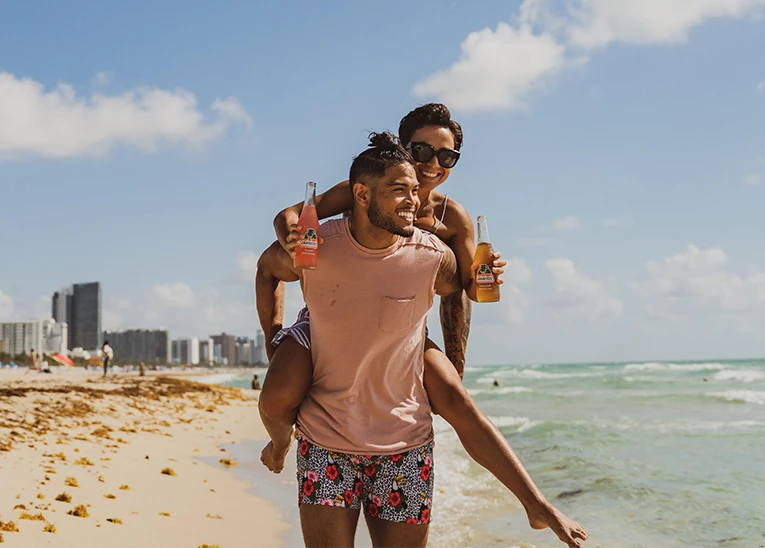 girl on the piggy-backing on a guy as he walks down the beach and she holds onto two jarritos