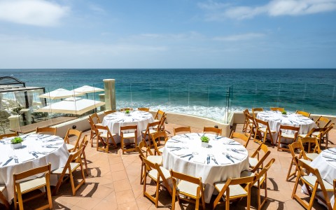 a table set up with white cloths and chairs on a patio overlooking the ocean