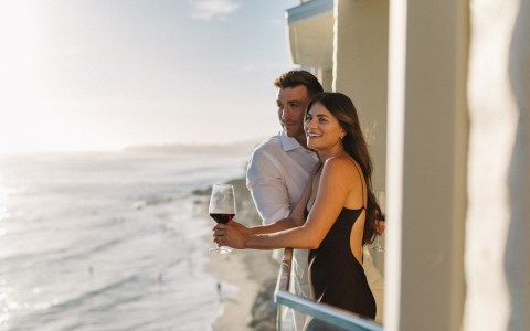 a man and woman holding wine glasses on a balcony overlooking the ocean