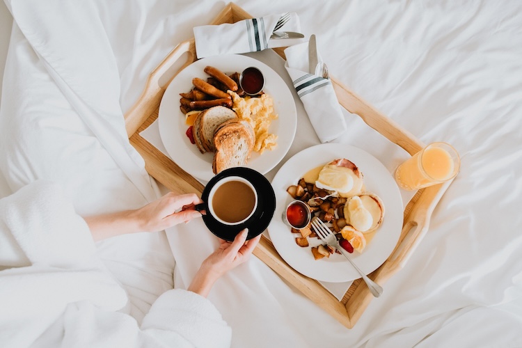 a person holding a cup of coffee next to plates of food on a tray in bed wearing a white robe