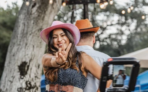 a woman in a dress and hat making a peace sign with a man behind her