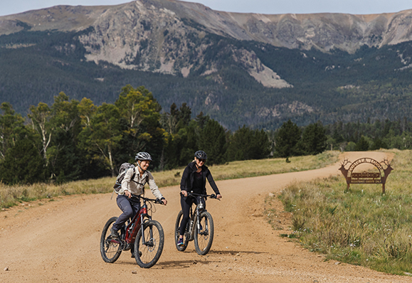 a couple of people riding bikes on a dirt road with mountains in the background