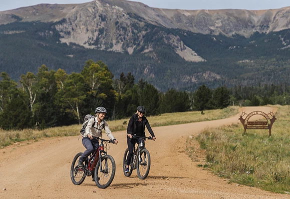 a couple of people riding bikes on a dirt road with mountains in the background