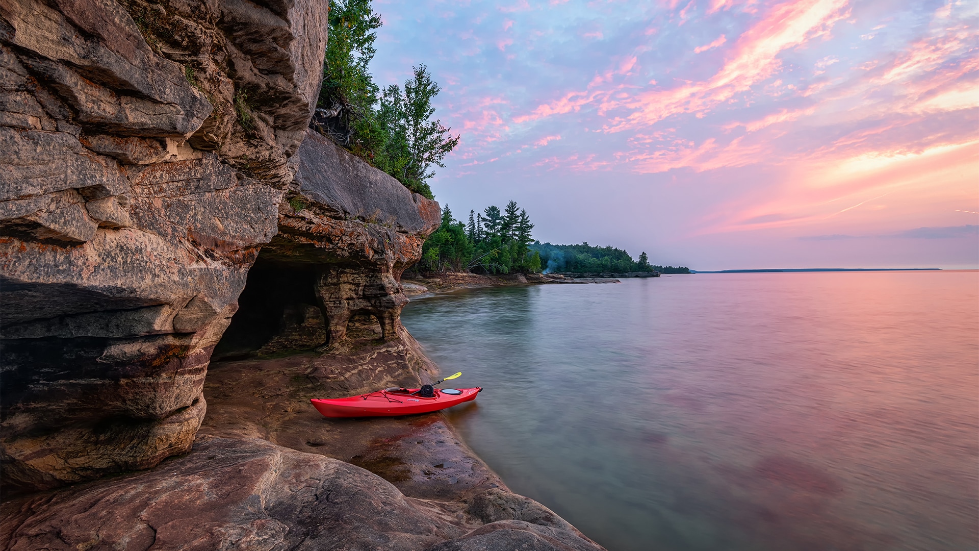 a red kayak on a rocky shore with trees and a pink sky
