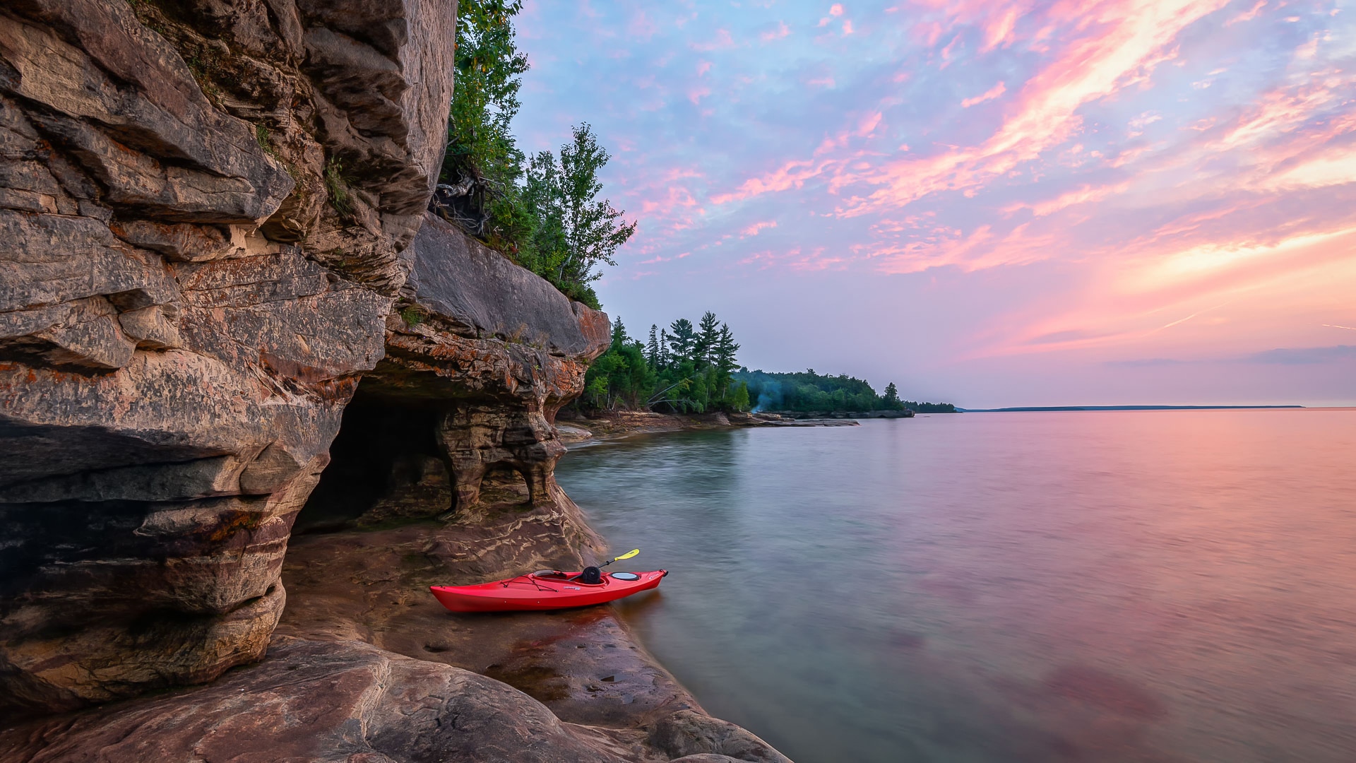 a red kayak on a rocky shore with a cave and trees