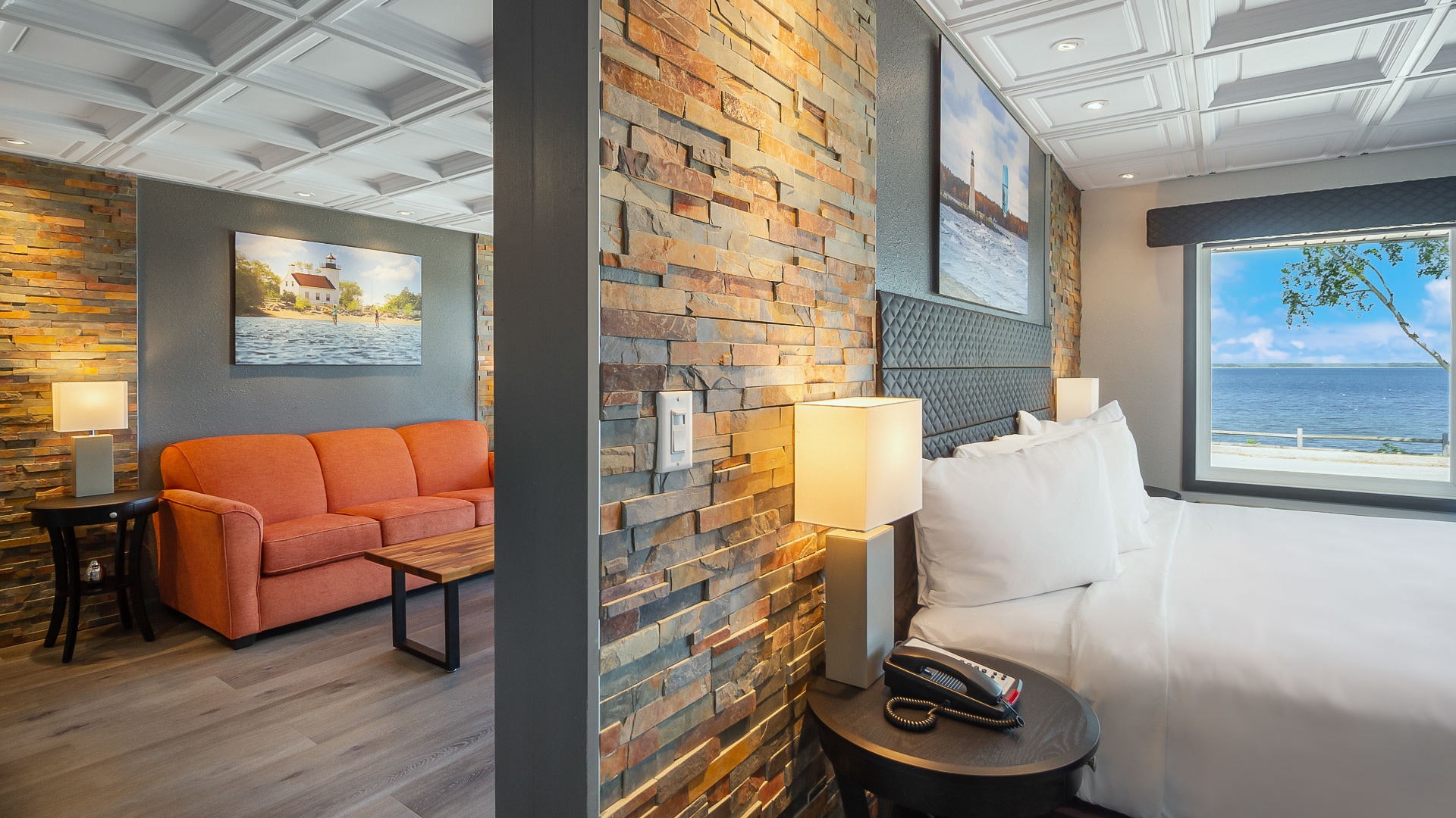 Modern hotel suite with living and bedroom areas, stone walls, and a water view