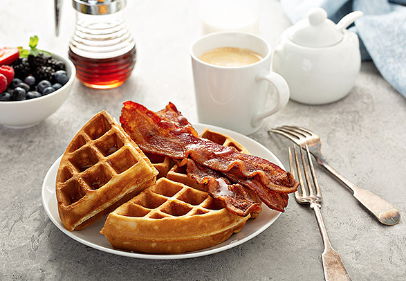 a waffle with bacon on top with a cup of coffee by the side and a bowl with fruit