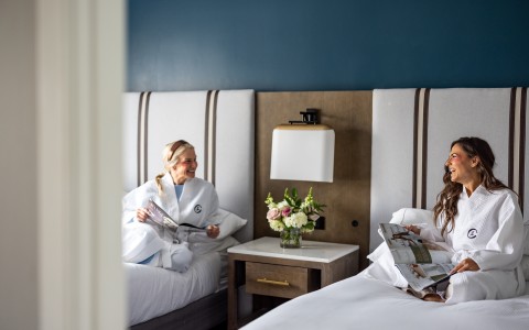 a woman in white robe reading a magazine while another woman is lying on the bed