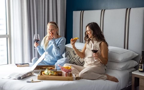 a group of women sitting on a bed eating pizza and drinking wine