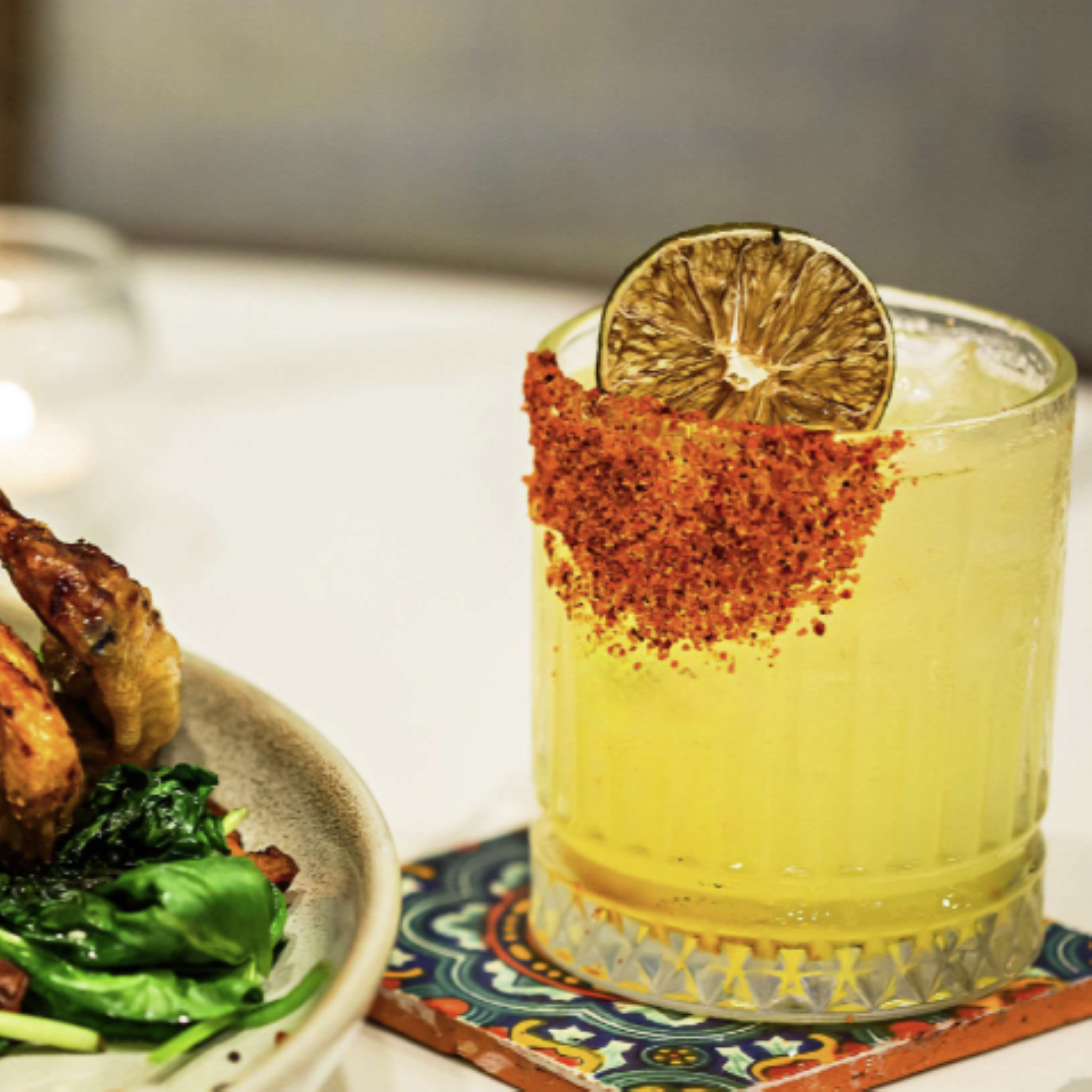 a glass of yellow liquid with a lime slice on top of a plate of food