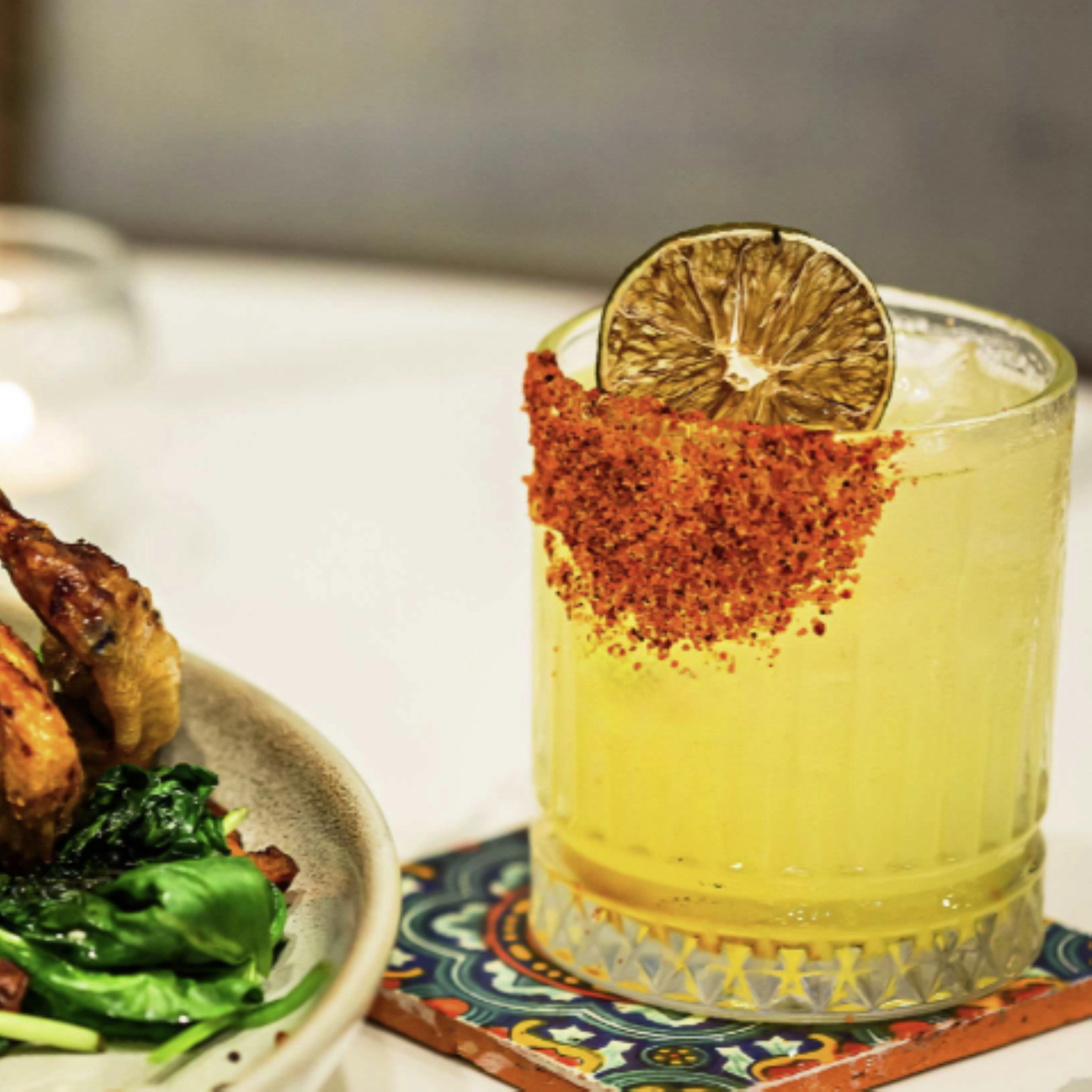 a glass of yellow liquid with a lime slice on top of a plate of food