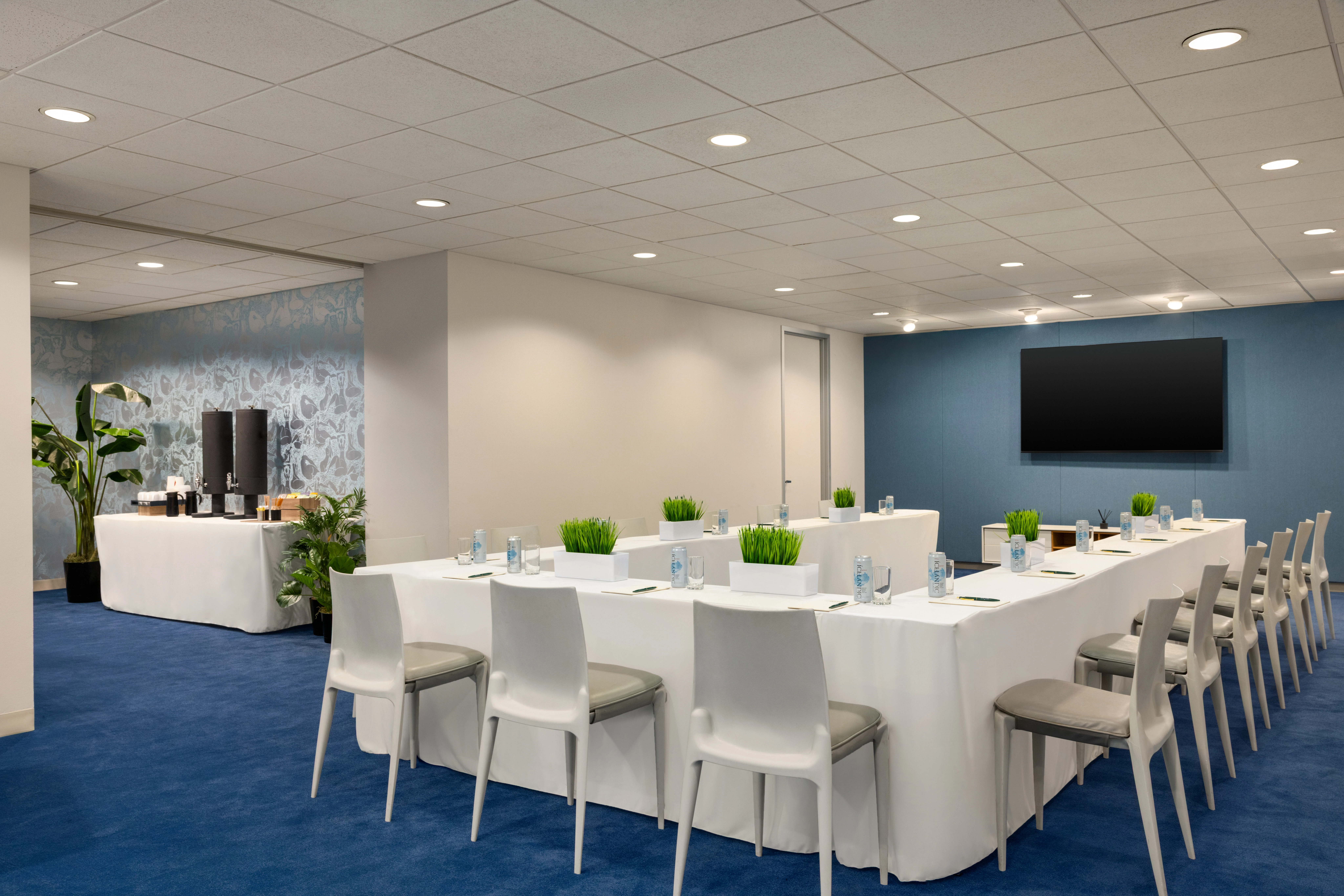 A meeting room with a long white table, white chairs, green plants, and a large screen on the wall.