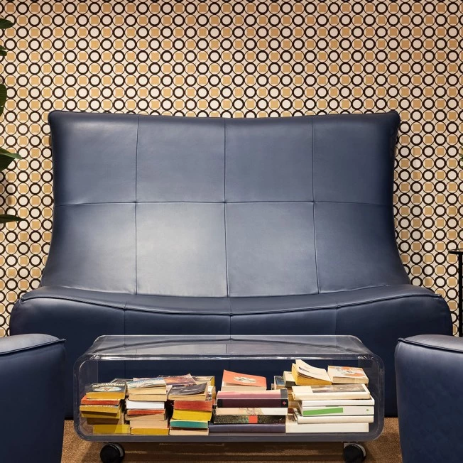 A blue leather sofa and two ottomans with a glass coffee table filled with books in front of a patterned wallpaper.