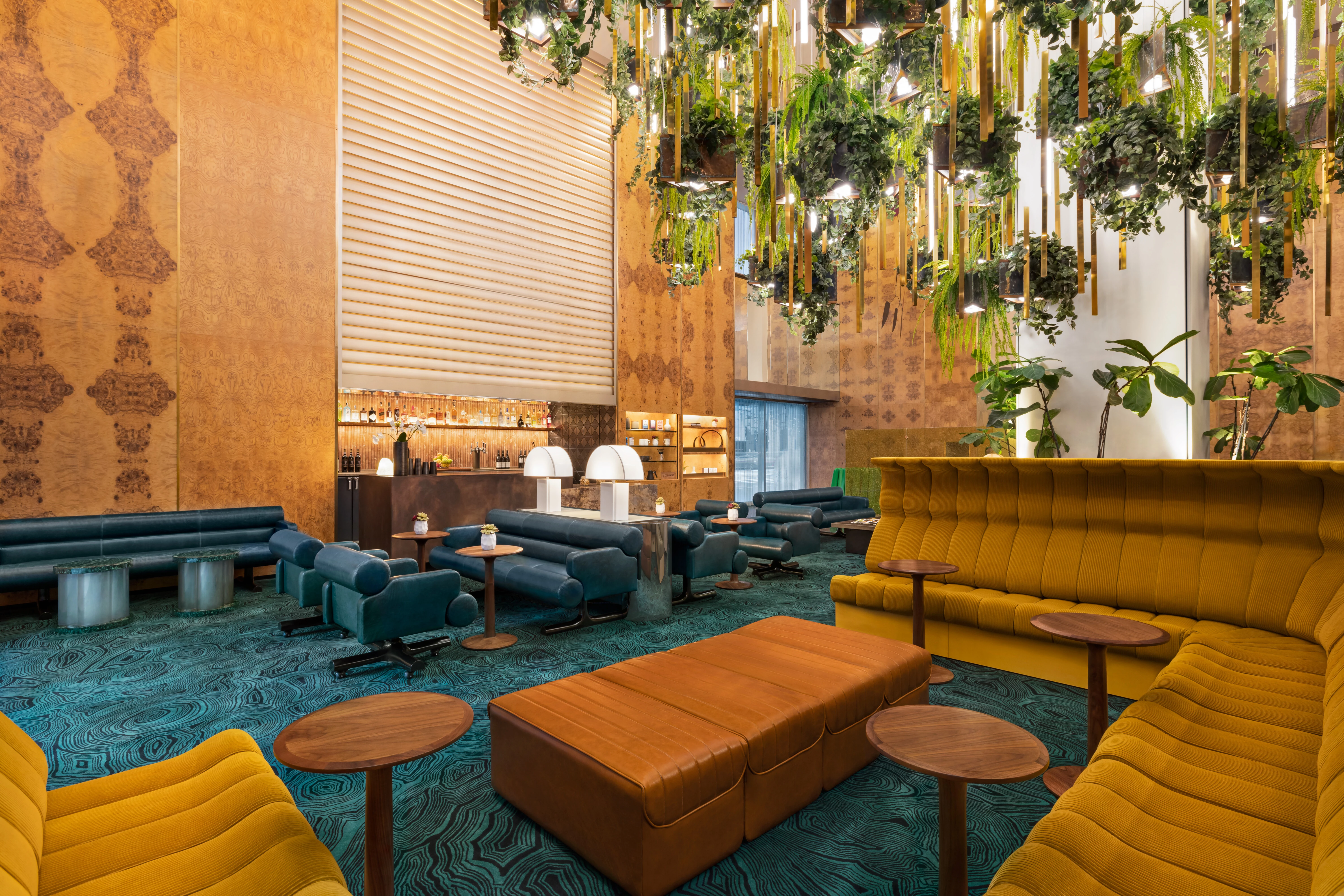 A wide view of a hotel lobby with yellow and blue sofas, wooden tables, and hanging plants.