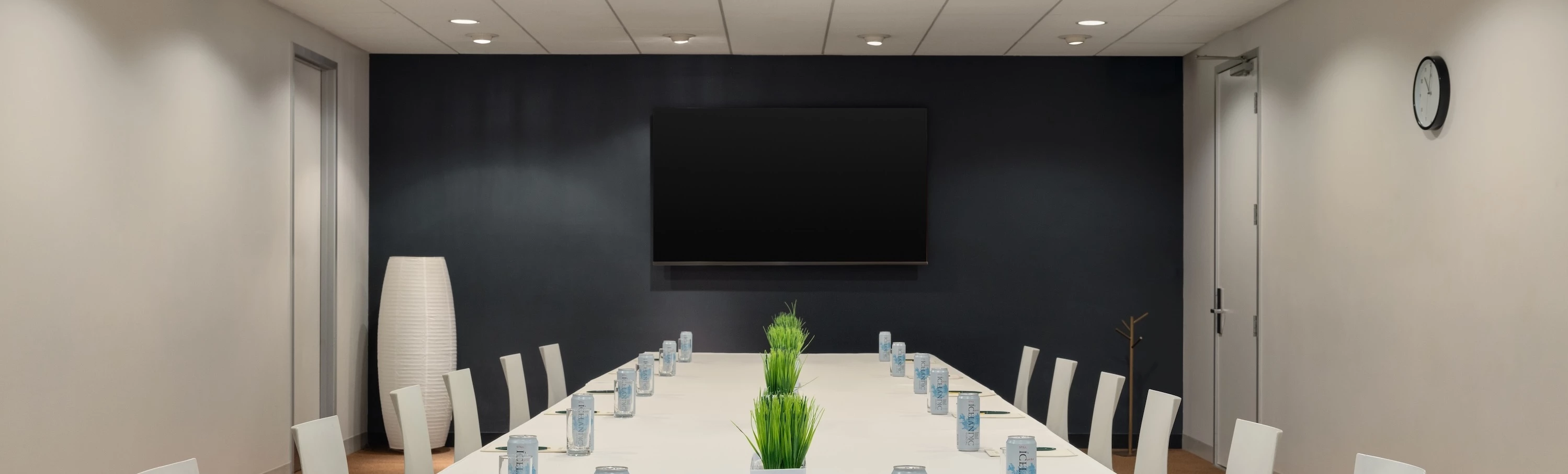 A conference room with a long white table, white chairs, a large TV screen on a dark wall, and plants in the center of the table.