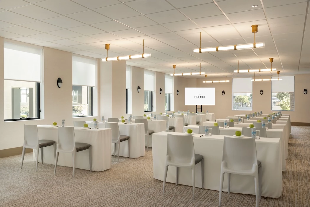 A well-lit meeting room with white tables and chairs, a projector screen, and windows.