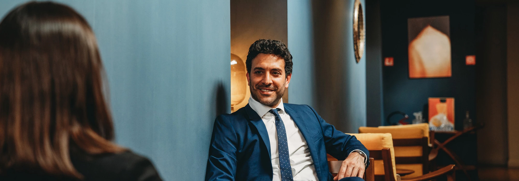 A man in a blue suit sitting in a lobby lounge with blue walls and yellow chairs.