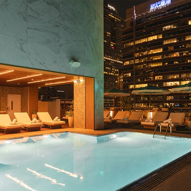 A nighttime view of a rooftop pool with lounge chairs and umbrellas, overlooking a cityscape.