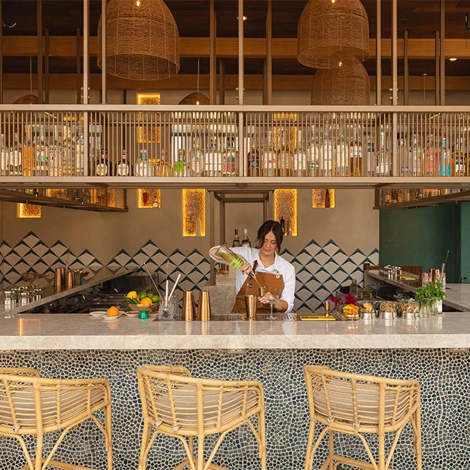 A bartender prepares a drink at a bar with a tiled front, stools, and a counter.