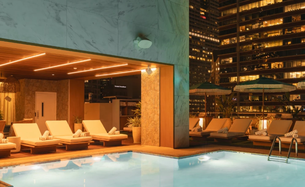 A nighttime view of a hotel pool with lounge chairs and city skyline.