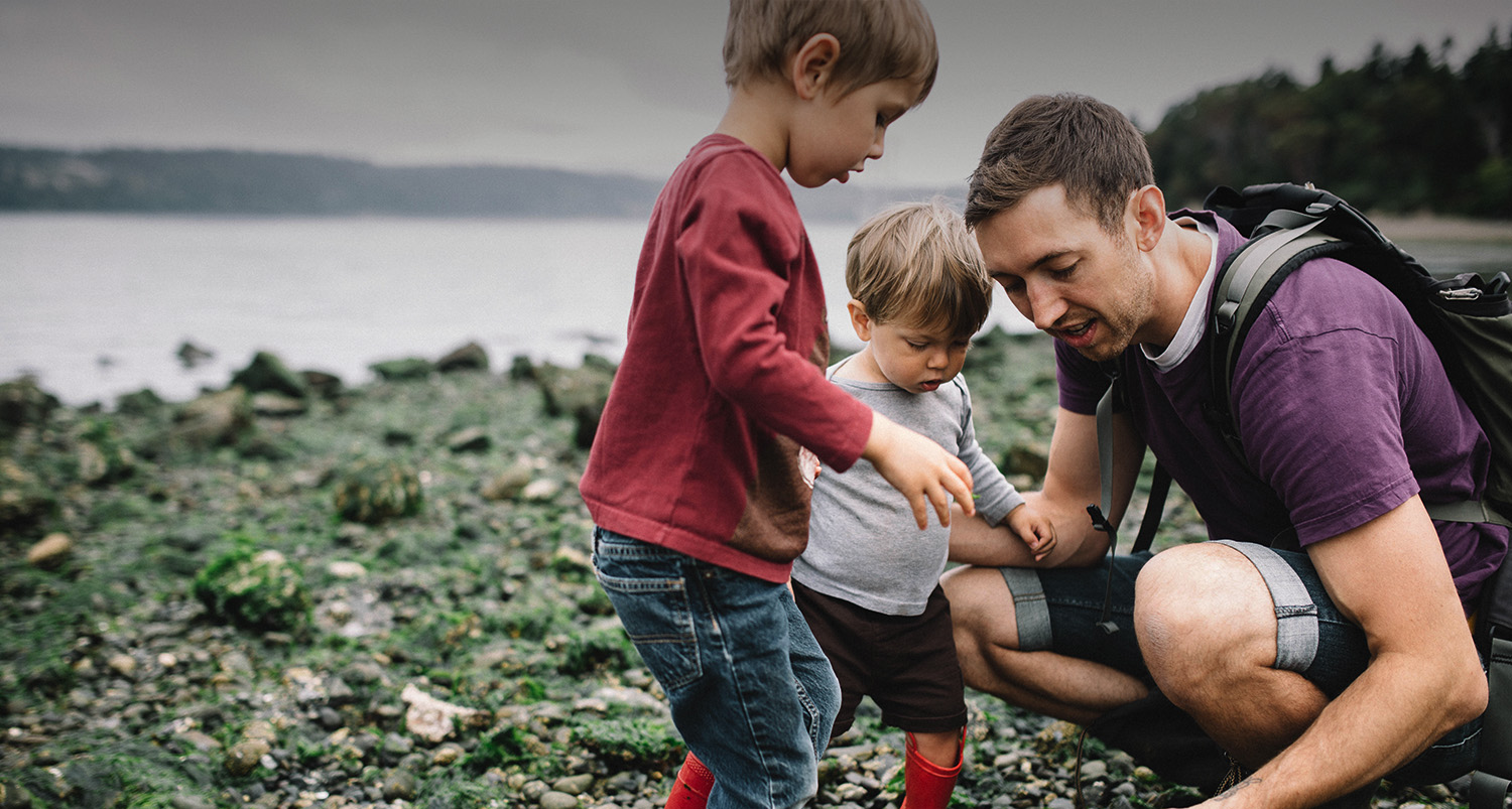 A man kneeling down looking a pebbles with two little boys near the water.