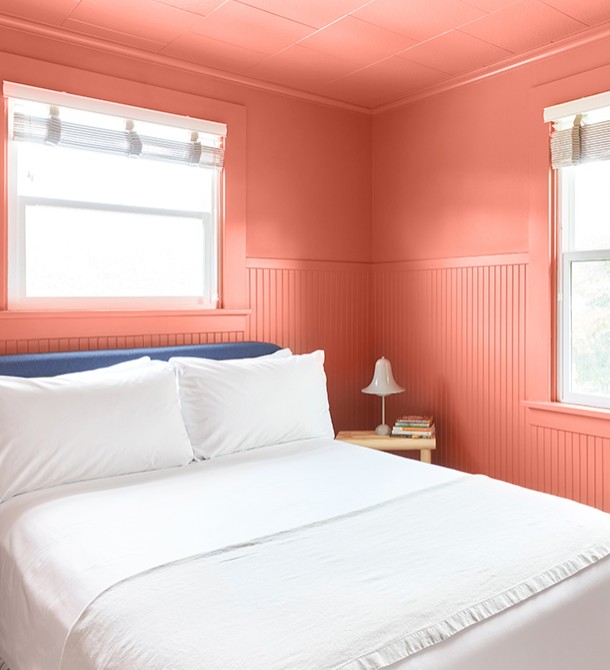 A light pink room with a bed and a small table and the sun shining in from the windows.