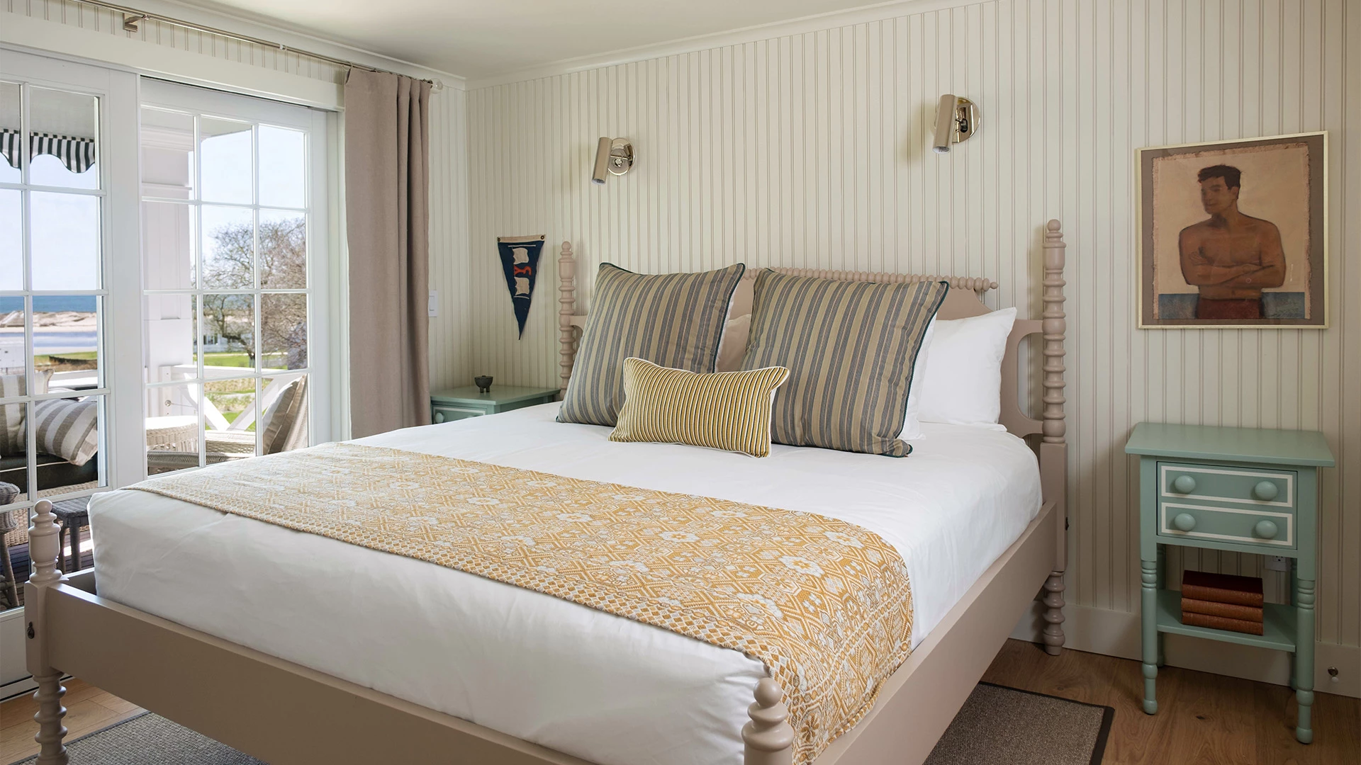 A neatly made bed with pillows and a yellow blanket in a room with white walls, a window, and a painting.