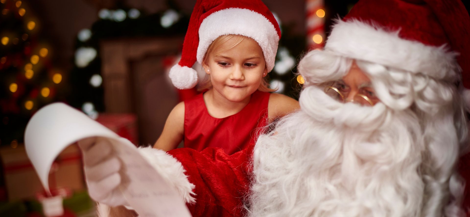 a girl in a red dress and hat looking at a santa claus