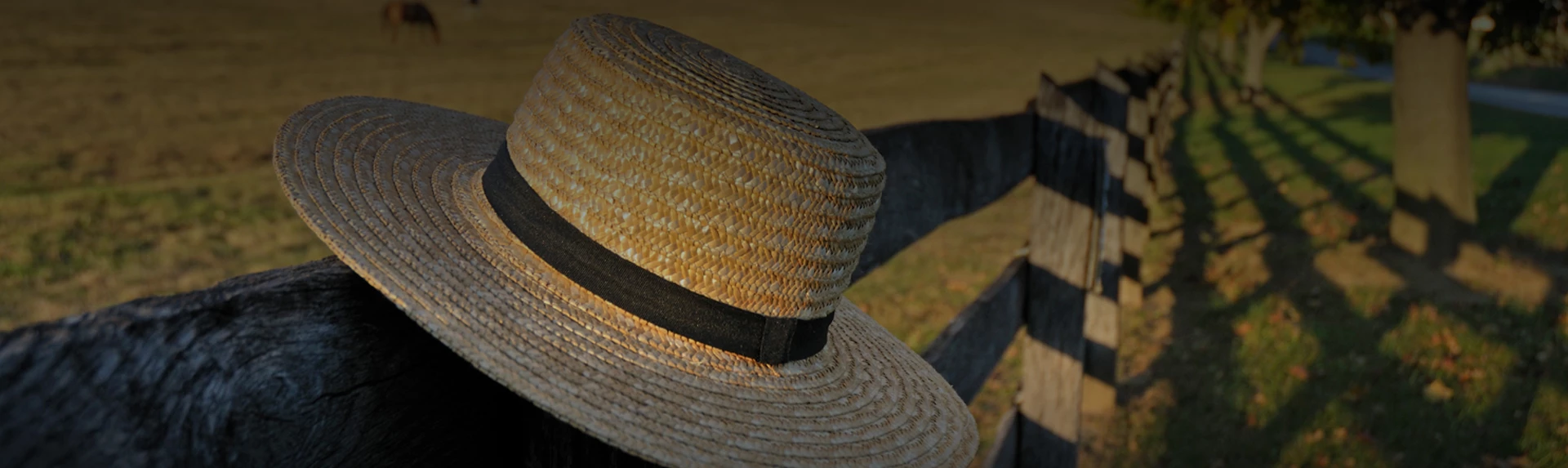 A dim lit photo of a round straw hat hanging on a wooden fence post.