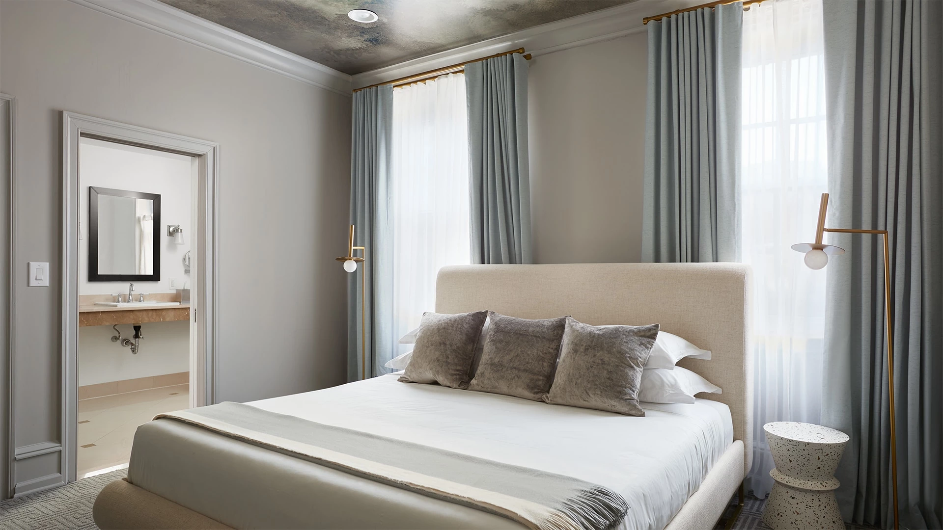 A hotel room with a bed, window, curtains, lamps, and a doorway to the bathroom