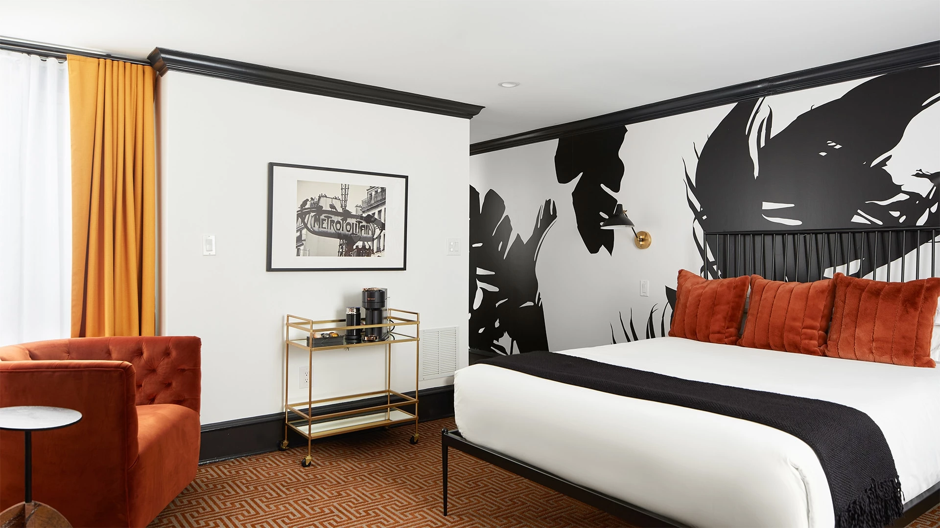 A hotel room with a bed, chair, and cart in front of a window with orange curtains.