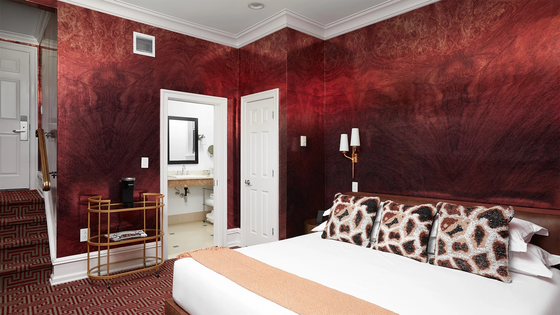 A wide view of a hotel room with a red accent wall, white bed, and gold furniture.