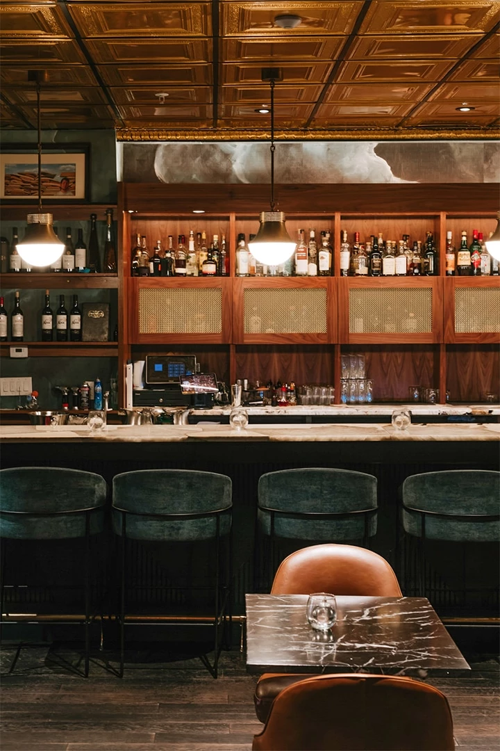 A dimly lit bar with a marble countertop, velvet chairs, and wooden shelves stocked with glass bottles, under pendant lights.