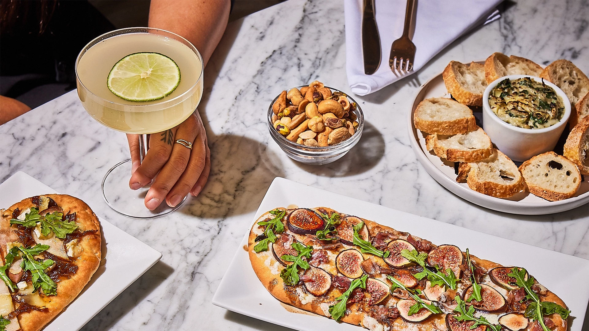 A table setting with a cocktail, flatbread, nuts, and bread on a marble table.