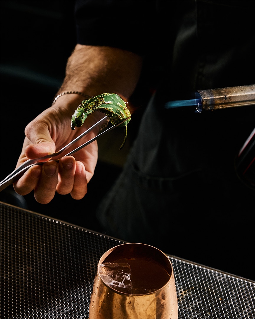 A bartender is preparing a drink with a green herb and a torch.