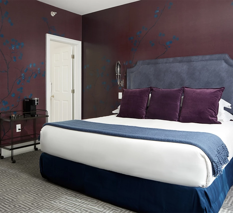 A hotel room with a king-sized bed, purple accents, and a blue headboard.