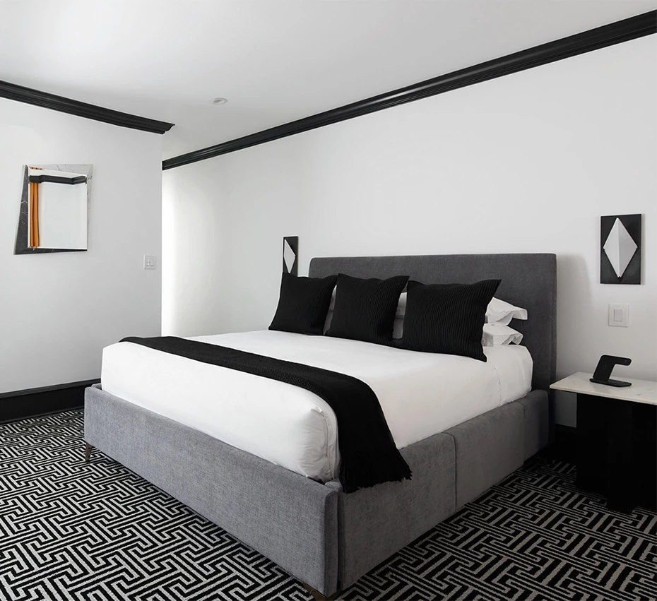 A wide view of a modern hotel room with a large bed, nightstand, and geometric carpet.