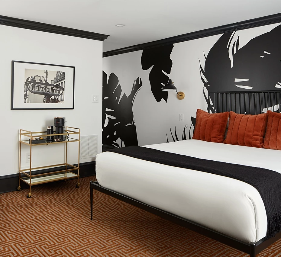 A large bed with white bedding and orange pillows is centered in a room with a black and white leaf-patterned wall, a gold bar cart, and a framed picture.