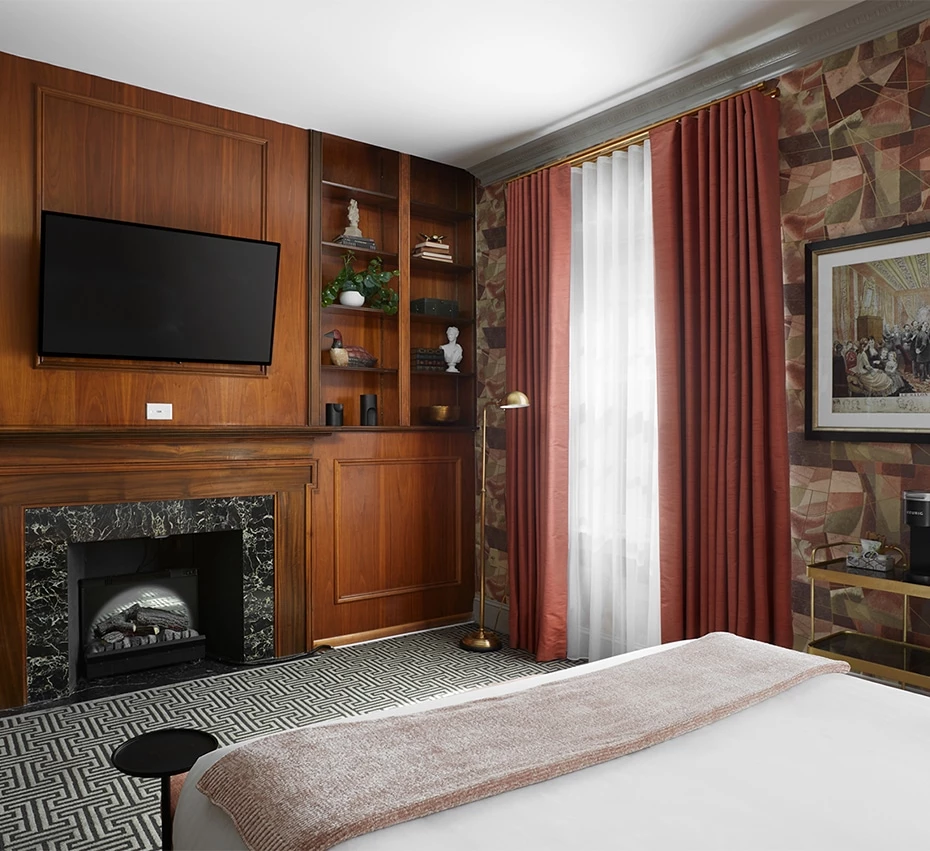 A luxurious hotel room with a large bed, marble fireplace, flat-screen TV, and patterned carpet.