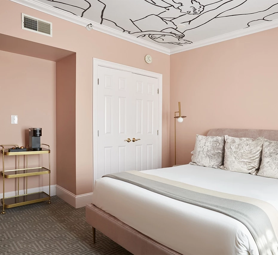 A wide view of a hotel room with a king-sized bed, pink walls, a white door, and a ceiling mural.