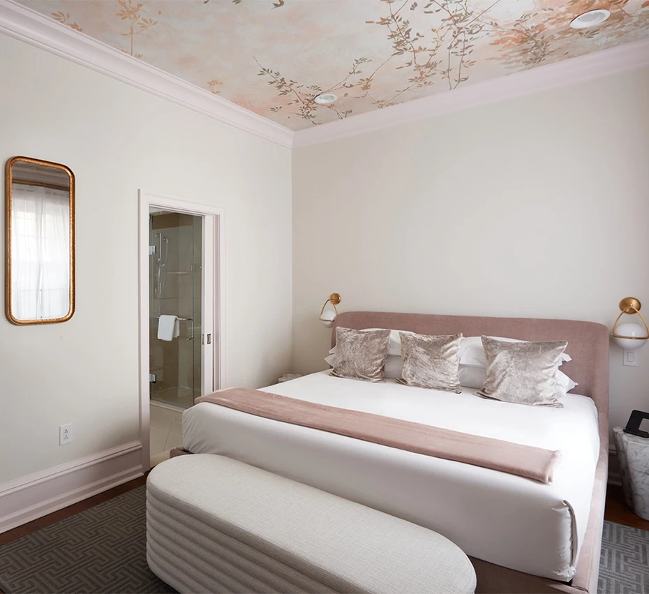 A wide view of a hotel room with a pink headboard, white bedding, and a floral ceiling mural.