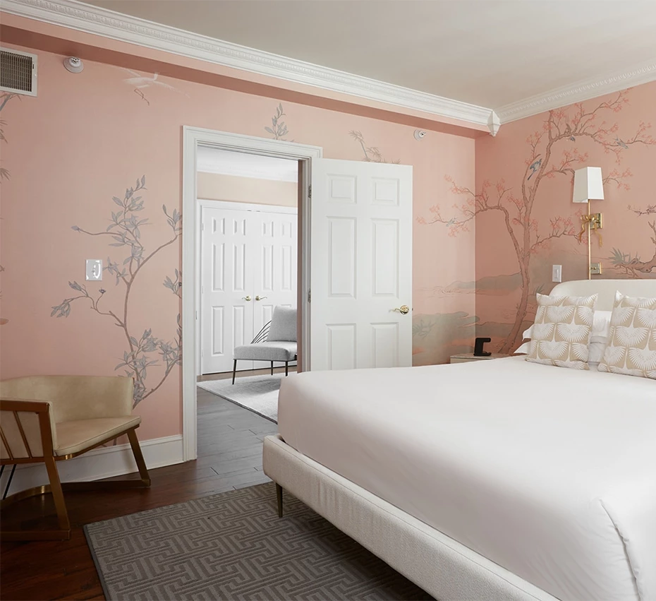 A wide view of a hotel room with a pink floral wallpaper, a white bed, and a chair.