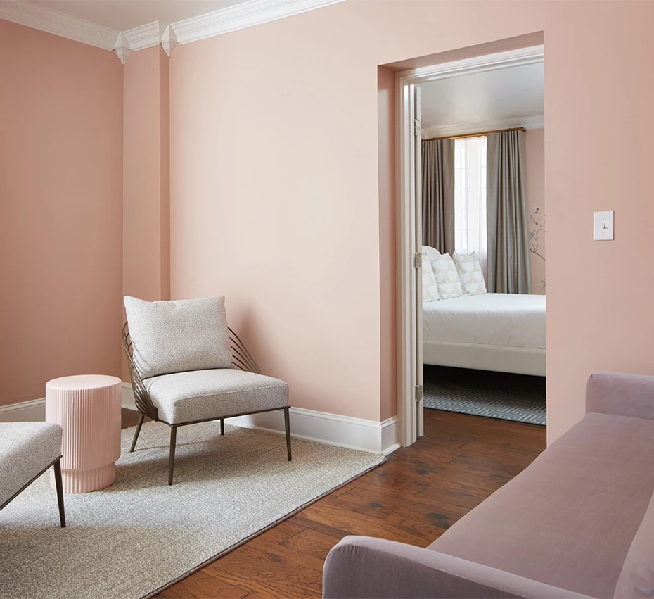 A wide view of a blush-pink room with an open doorway leading to a bedroom with a bed.
