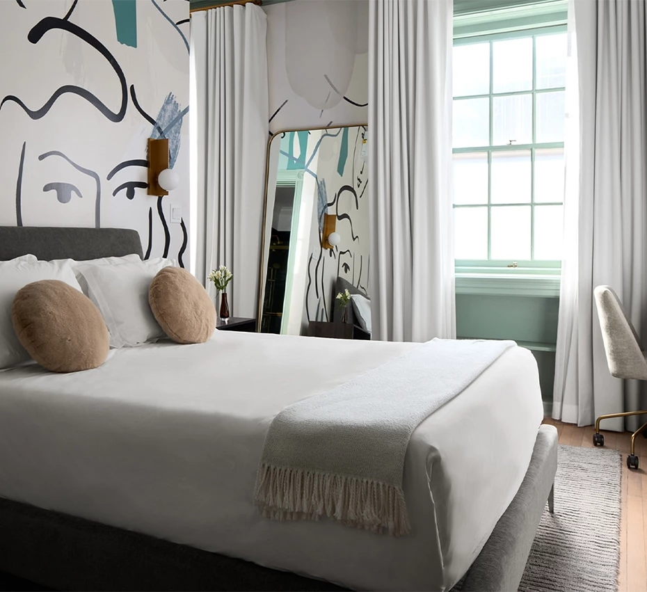 A neatly made bed with white linens and a gray upholstered footboard, with a large window, abstract art on the wall, and a chair in the corner.